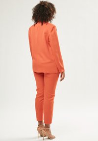 Oranges Anzugset bestehend aus einem taillierten Blazer und schlank geschnittenen Hosen. Der Stoff wirkt glatt mit einem leichten Glanz und wird durch minimalistische Designelemente akzentuiert.