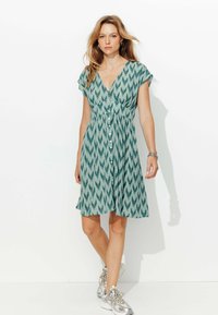Robe verte à motifs avec un décolleté en V, des manches courtes et une fermeture à boutons. Le tissu texturé présente un motif en zigzag.