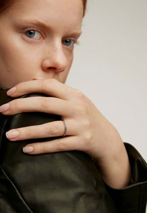 Anillo de plata con una banda texturizada, llevado en una mano con uñas de color rosa pálido, descansando sobre una chaqueta de cuero negra. Fondo neutro.