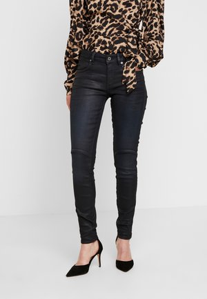 Femme portant un jean skinny noir, un chemisier à manches longues imprimé léopard, et des chaussures à talons noirs pointues, debout devant un fond uni.