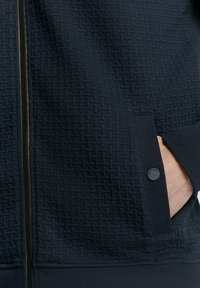 Donkerblauwe zip-up jas met een textuurpatroon, zijzakken met matte zwarte knopen en een contrasterende ritsdetail.