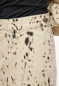 Pantalon beige à surface texturée, avec des motifs éclaboussés noirs et un bouton bronze à la ceinture.