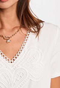 Top blanco con ribete de encaje circular en el escote en V, detalle de bordado floral texturizado, combinado con un collar de cadena dorada con charm y acento de perla.