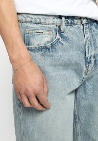 Helle blaue Jeans aus Denim mit einer distressed Optik, ausgestattet mit einer Vordertasche und einem silbernen Armband am Handgelenk, das auf dem Oberschenkel ruht.