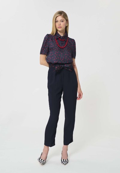 Femme blonde debout portant un chemisier bleu marine à motifs avec des perles rouges, un pantalon taille nouée bleu marine, et des escarpins à talons rayés noir et blanc.