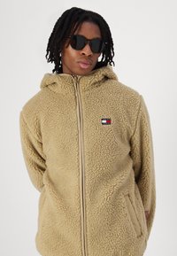 Beige Fleece-Hoodie mit Struktur, Reißverschluss und Seitentaschen. Mit kleinem rot-, weiß- und blauem Markenlogo auf der Brust. Dunkle Sonnenbrille getragen.