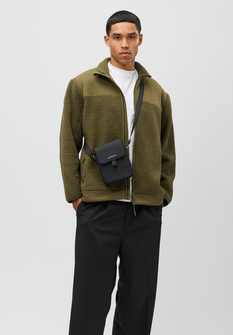 Olivgrön fleecjacka med dragkedja och en svart axelväskan. Modellen bär svarta byxor och en vit t-shirt med rund halsringning. Enkla designfunktioner.