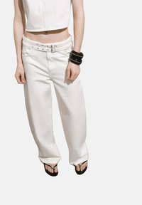 Pantaloni bianchi a gamba larga con una finitura texturizzata, caratterizzati da una vita alta, dettagli con doppia fibbia e tasche laterali. Indossati con ciabatte nere.