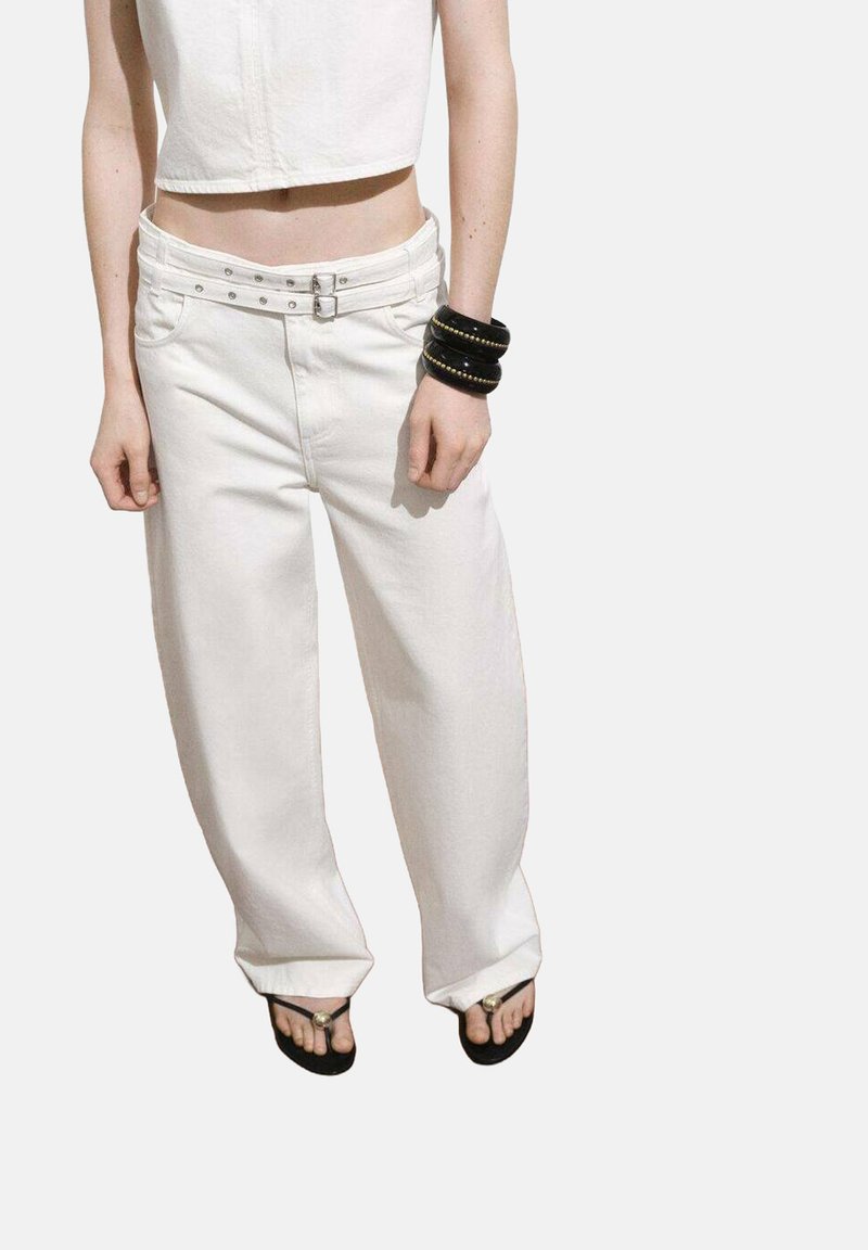 Pantaloni bianchi a gamba larga con una finitura texturizzata, caratterizzati da una vita alta, dettagli con doppia fibbia e tasche laterali. Indossati con ciabatte nere.