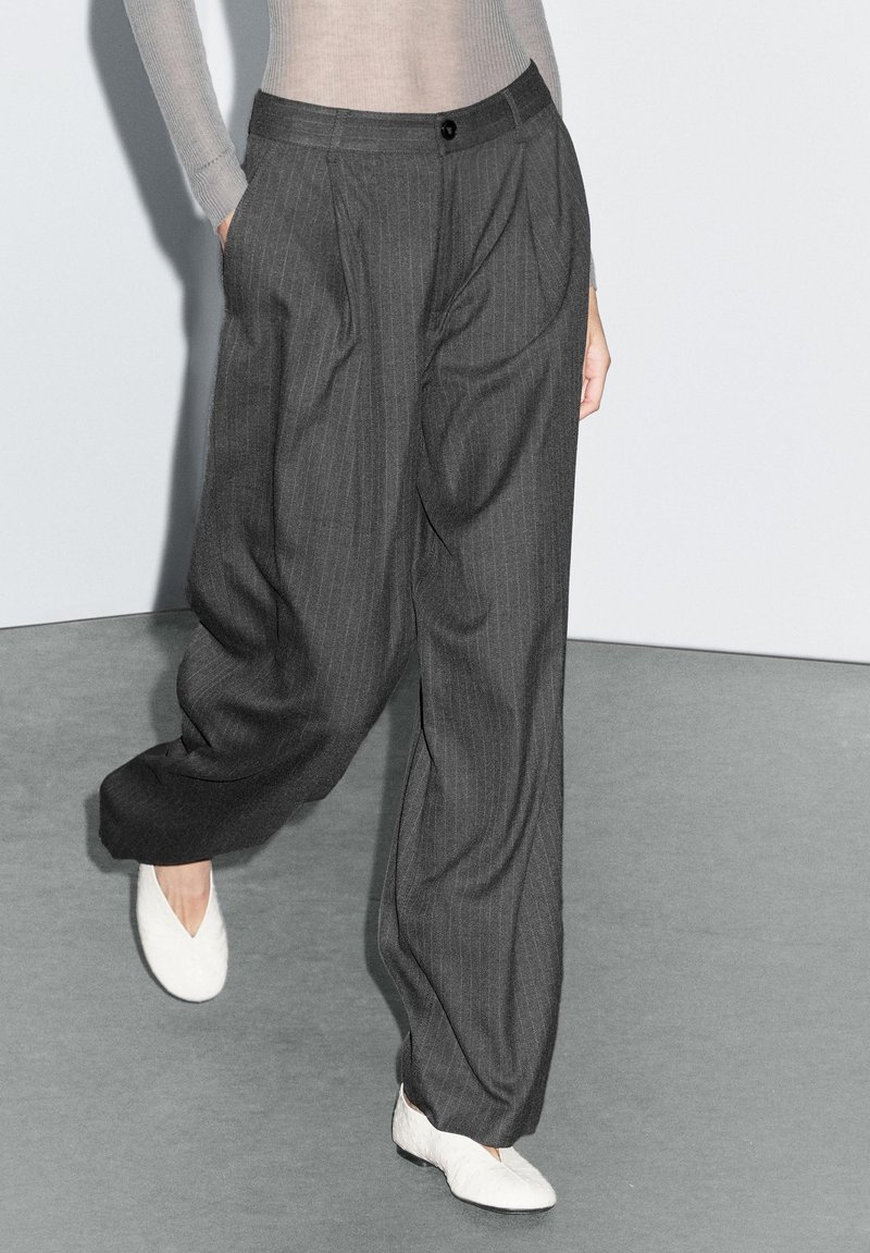 Dark grey pinstriped trousers with a relaxed fit, featuring side pockets and a front button closure, paired with white textured shoes.