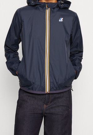 Navy blue hooded jacket with yellow, white, and blue striped zipper and hands in pockets, worn with dark denim jeans.