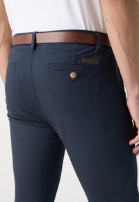 Pantalones azul marino de mezcla de algodón, con cinturón de cuero marrón, dos bolsillos traseros con botones y una etiqueta con el logo en la cintura.