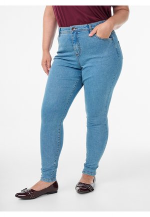 Personne portant un jean skinny bleu clair et des ballerines bordeaux foncé avec un détail de nœud, debout avec une main dans la poche sur fond blanc.