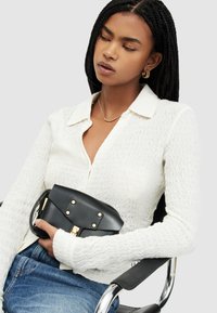 Top bianco in maglia strutturata con colletto, abbinato a jeans blu. La modella tiene una piccola borsa nera con hardware dorato e borchie.