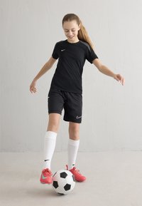 Must musta sportlik särk ja lühikesed püksid valge Nike logoga, valged põlvikud ja roosad jalgpallisaapad. Mustvalge jalgpall jalgade juures.