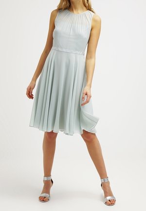 Robe sans manches bleu clair arrivant aux genoux avec une jupe plissée, portée avec des talons hauts argentés à bride cheville, femme debout les jambes écartées.