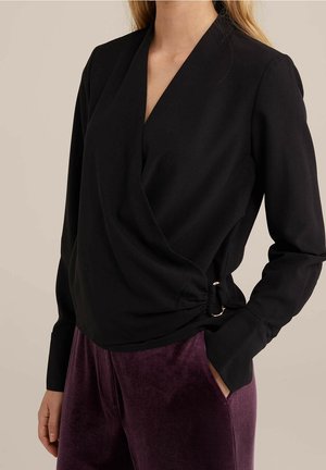 Vrouw draagt een zwarte wikkelstijl top met lange mouwen en een gesp aan de zijkant, en paarse fluwelen broek tegen een neutrale achtergrond.