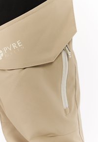 Beige broek gemaakt van lichte stof, met een ritszak en een opgevouwen tailleband waarop het logo "PVRE GREEN" prominent is weergegeven.