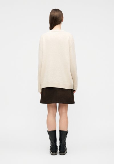 Γυναίκα με μακριά καστανά μαλλιά που φοράει εκρού φαρδιά πουλόβερ, σκούρα καφέ φούστα και μαύρες μπότες μέχρι τη μέση της γάμπας, στέκεται γυρισμένη από την πλάτη σε λευκό φόντο.