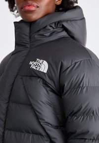 Svart boblejakke laget av glatt nylon med høy krage, med quiltede seksjoner og en fremtredende hvit logo på brystet.