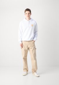 Felpa bianca con cappuccio e logo "HUGO" arancione, abbinata a pantaloni cargo beige chiaro. Le sneakers sono bianche con lacci bianchi. Sfondo minimalista.