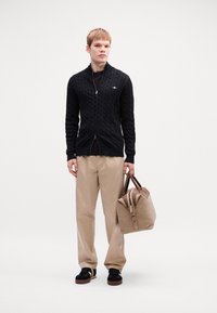 Fekete kötött kardigán, elöl cipzárral, bézs széles nadrággal, fekete cipővel és világosbarna bőrtáskával, barna részletekkel. A modell középen áll.