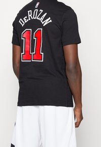 Camiseta de algodón negra con 'DE ROZAN' y el número '11' en rojo con contorno blanco, y etiqueta del logo de la NBA en la parte posterior. Mangas cortas, corte estándar.