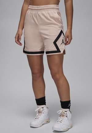 Persoon die beige basketbalshorts draagt met zwarte bies en logo, zwarte sokken en witte hoge sneakers, staand tegen een grijze achtergrond.