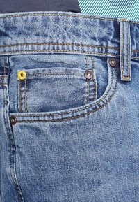 Primer plano de unos vaqueros de denim azul que muestran el bolsillo frontal con remaches metálicos y costuras naranjas.