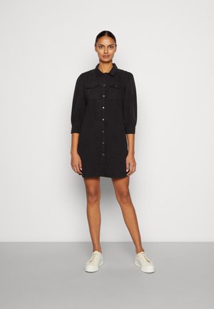 Zwart shirtjurk met knoopsluiting, twee borstzakken en driekwartmouwen. Het combineren met witte sneakers versterkt de look.