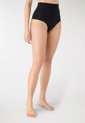 Donna con slip modellante a vita alta nero, in piedi scalza contro uno sfondo bianco semplice.