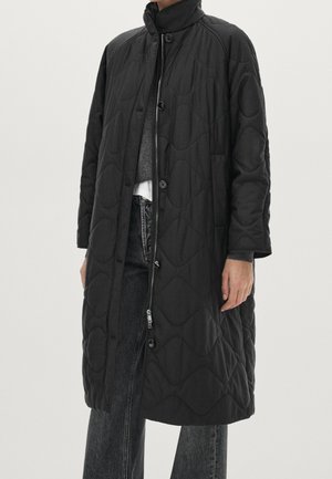 Long manteau noir matelassé avec col montant, fermeture par boutons et fermeture éclair, porté sur un pull gris et un jean noir.