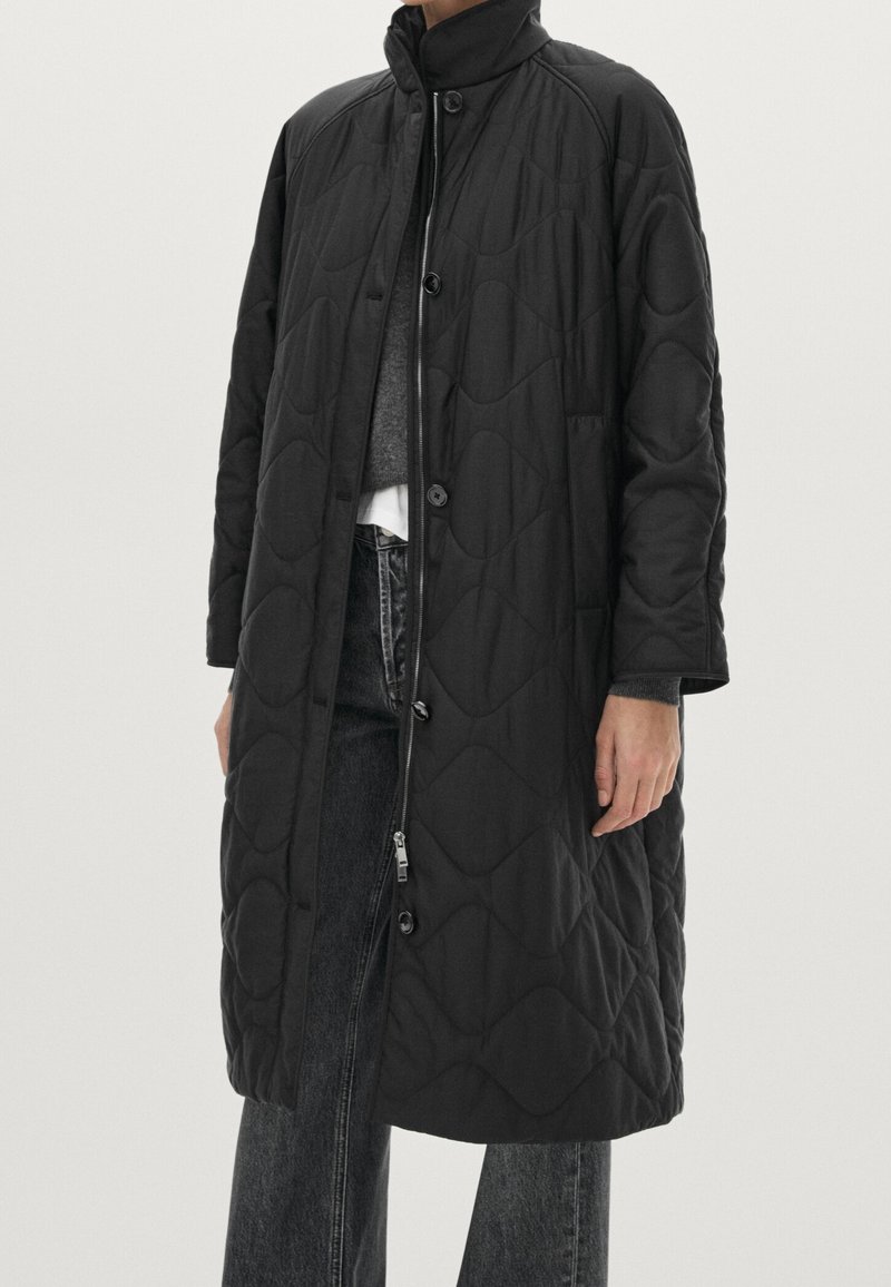 Long manteau noir matelassé avec col montant, fermeture par boutons et fermeture éclair, porté sur un pull gris et un jean noir.