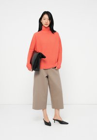 Kvinne iført korallfarget genser med høy hals, beige vide, forkortede bukser, svarte spissede hæler, og som holder en stor svart clutch-veske.