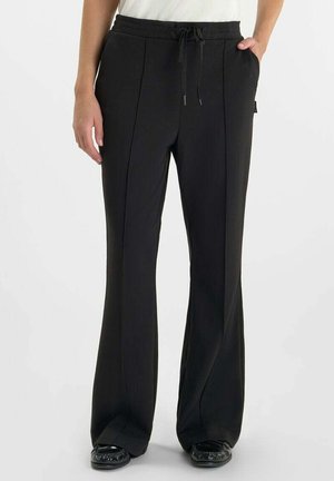 Persona que lleva pantalones negros de pierna ancha con costuras frontales y cintura con cordón, combinados con zapatos negros sobre un fondo liso.
