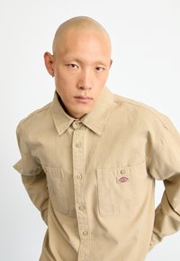 Camisa de botones color caqui, material de algodón, dos bolsillos en el pecho, parche con logotipo en el bolsillo izquierdo, corte relajado, cuello con solapa, mangas largas.