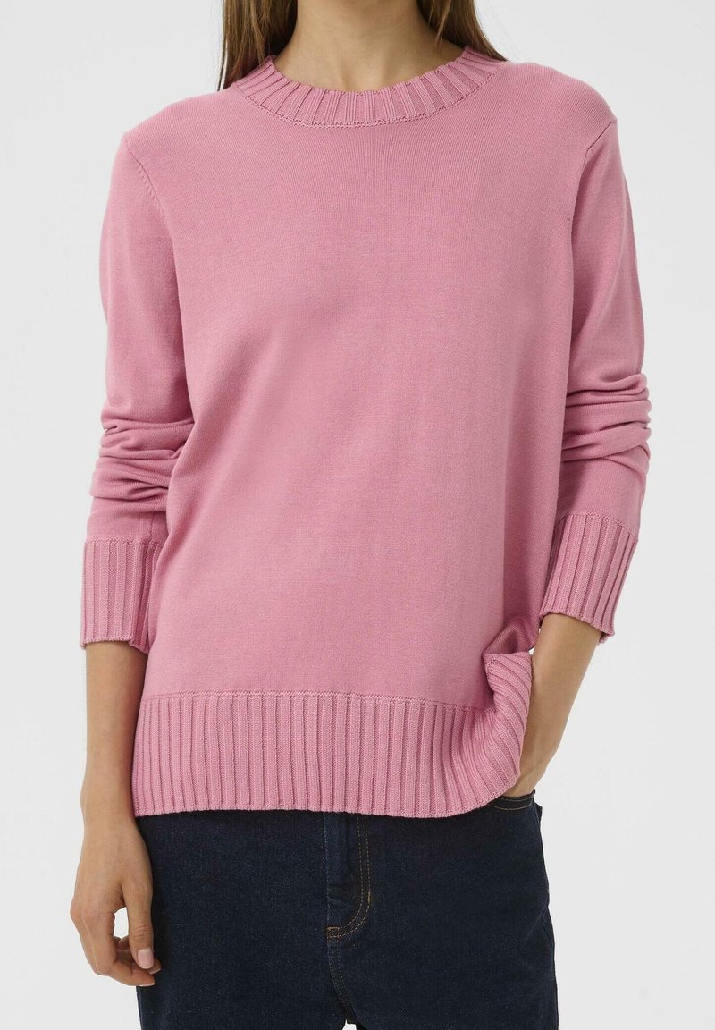Femme portant un pull rose uni avec poignets et ourlet côtelés, une main dans la poche d’un jean bleu foncé.