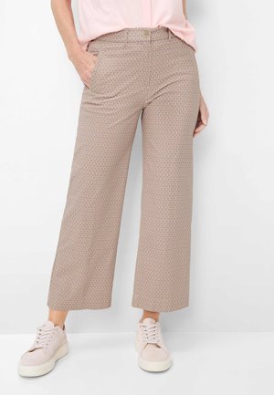 Vrouw draagt beige wijde cropped broek met patroon en zijzakken, en lichtroze sneakers, staand tegen een witte achtergrond.