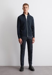 Marine blauw ritsvest met hoge kraag, voorzien van een textuur breisel, gecombineerd met slim-fit donkere broek en zwarte instappers.