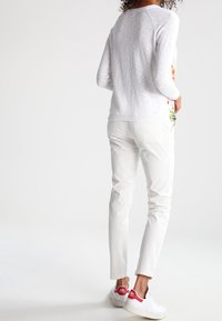 Frau, die einen weißen Pullover, weiße Hosen und weiße Sneaker mit roten Akzenten trägt, steht vor einem schlichten weißen Hintergrund, mit dem Rücken zur Kamera.