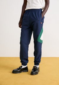 Mörkblå träningsoveraller med elastisk midja, sidofickor och gröna samt vita geometriska accenter. Matchat med svarta skor och vita strumpor.