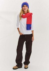 Niebiesko-czerwona dziana czapka zestawiona z pasującym czerwono-niebieskim szalikiem. Biała koszula z długim rękawem, brązowe spodnie o szerokich nogawkach oraz beżowe buty.