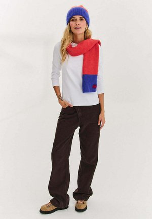 Niebiesko-czerwona dziana czapka zestawiona z pasującym czerwono-niebieskim szalikiem. Biała koszula z długim rękawem, brązowe spodnie o szerokich nogawkach oraz beżowe buty.