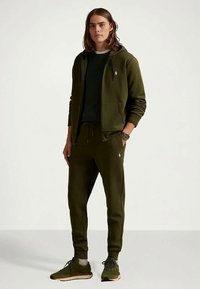 Jovem com cabelo castanho até aos ombros, vestindo um hoodie de fecho verde escuro, calças de jogging combinando, camiseta preta e ténis verdes, de pé contra um fundo branco.