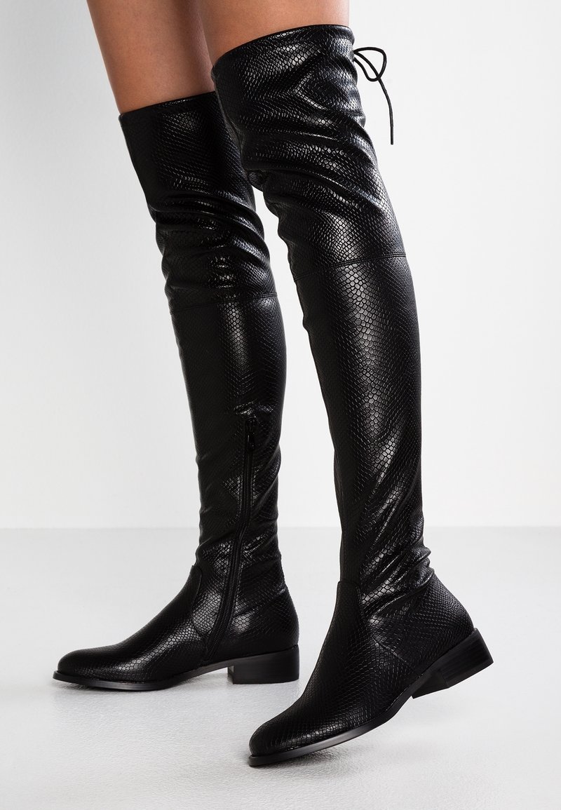 Botas de cuero negro, hasta la rodilla, texturizadas, con cremalleras laterales y cordones en la parte posterior, llevadas sobre piernas desnudas contra un fondo blanco.