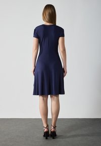 Lyhythihainen, polvenmittainen mekko tummansinisestä kankaasta, jossa on istuva yläosa ja levenevä hame; päällä mustat remmi-ökä high heel -kengät.