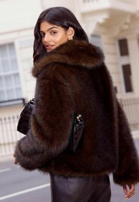 Ruda dirbtinio kailio striukė su dideliu apykakle, turinti tekstūruotą paviršių. Sukomponuota su juodu patentuotu rankiniu krepšiu su auksine sagtimi.