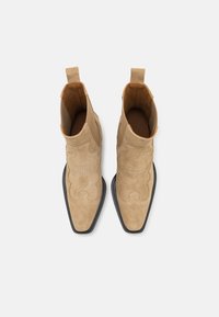 Botines de ante en color beige con puntera afilada, costuras decorativas y suela de goma negra. Cuenta con tiradores para facilitar su uso.
