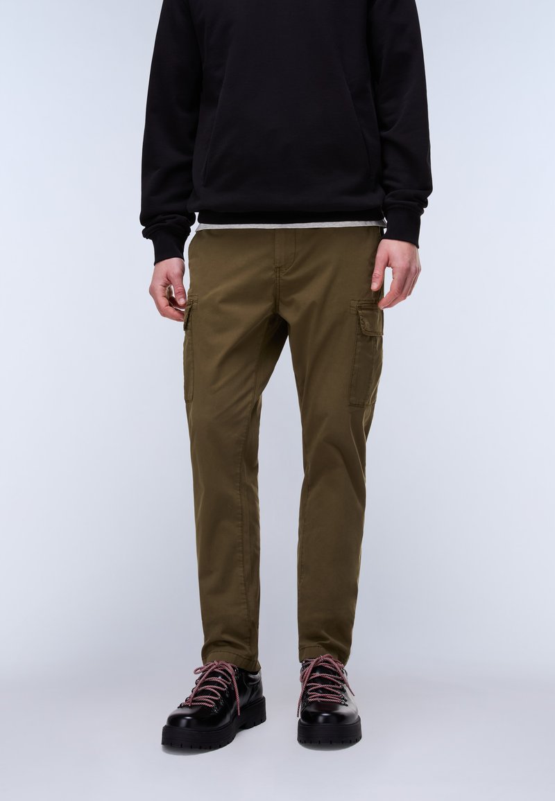 Person wearing olive green cargo pants, black sweater, and black leather boots with pink laces, standing against a plain light background.