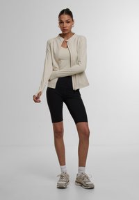 Lys beige lynlåsjakke over en tætsiddende cremet tanktop, parret med sorte cykelshorts. Modellen har grå sneakers og hvide sokker på.
